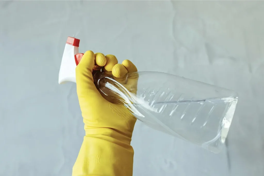Yellow gloved hand holding a clear spray bottle against a light background